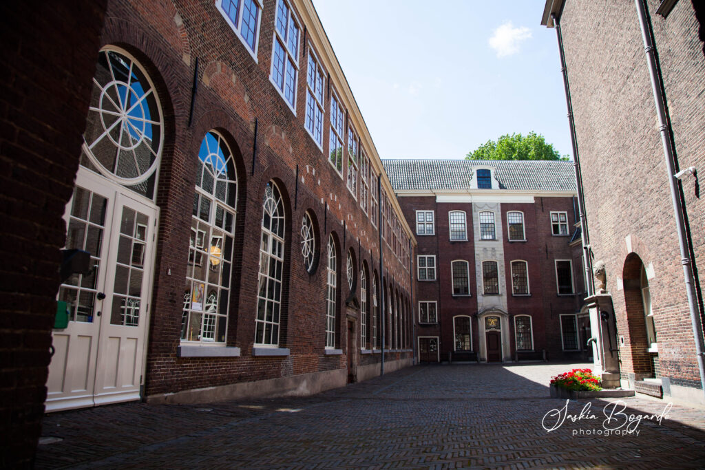 De binnenplaats van het Utopa Weeshuis in mei 2025. Links is er een groot bakstenen gebouw van twee verdiepingen met daarin witte klapdeuren en (half)ronde ramen met kleine ruitjes. Recht vooruit is een bakstenen gebouw te zien van drie verdiepingen met vergelijkbare ramen. Rechts is er alleen een hoekje van een bakstenen gebouw te zien, waar een stenen fontein uit komt met eronder rode bloemen. De binnenplaats zelf is bestraat met kasseien in een ruitpatroon. Fotograaf: Saskia Bogarde.