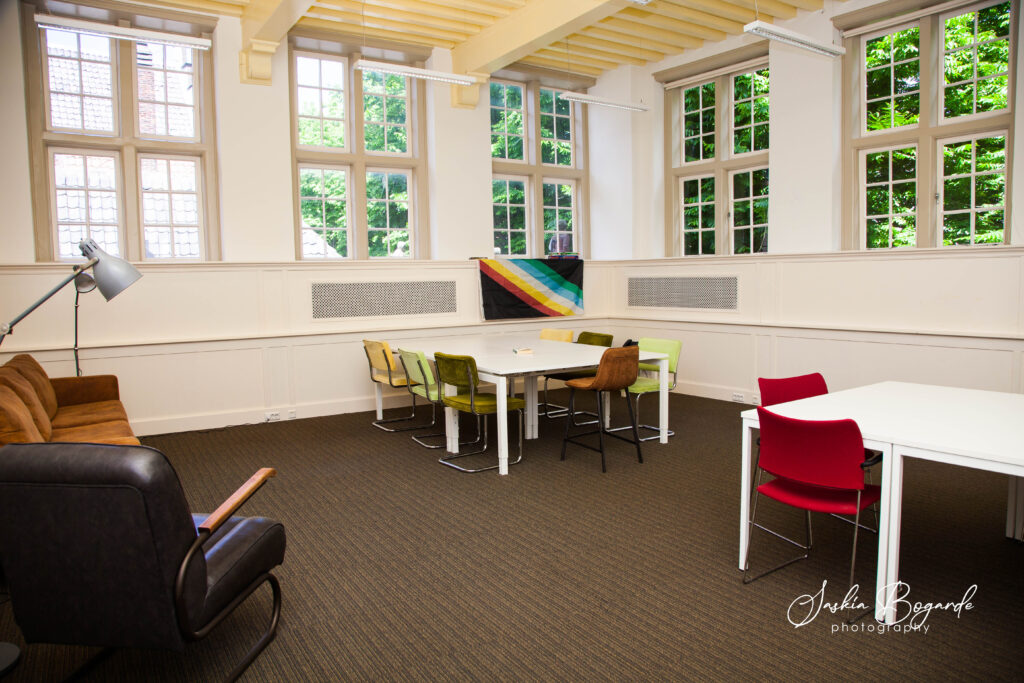 Het kantoor van het Kreukelarchief in mei 2025. Het is een lichte ruimte met een hoog plafond, met veel ramen met kleine ruitjes die uitzicht geven op grote bomen op de Hooglandse Kerkgracht. Links is een zithoek met fijne bank, fauteuil en stalampen. In de verre hoek van het kantoor is een vergadertafel met verschillende groene, gele en bruine stoelen. Erachter hangt de disability pride vlag aan de muur. Rechts staan wee witte bureaus tegen elkaar geschoven met rode stoelen eraan. Er ligt bruin tapijt met lange banen erin op de vloer. Fotograaf: Saskia Bogarde.