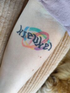 Een tatoeage op een arm met een witte huidskleur. Het woord 'Kreukel' staat met golvende letters op een achtergrond van speelse vegen in alle kleuren van de regenboog. Het ontwerp van de tatoeage is van Cee Burgundy. Op de achtergrond zijn een stukje roze mouw, een zacht bruin kussen en een hondenvacht te zien.