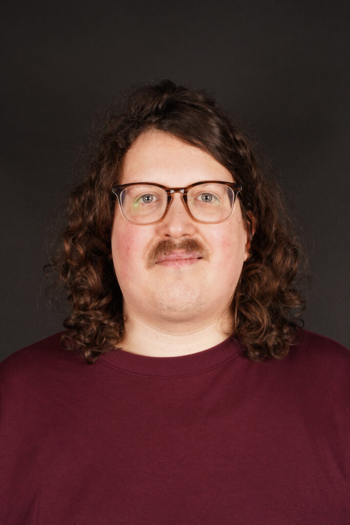 Portretfoto van Sem Bannenberg. Sem is een witte man van 35 met schouderlange, donkere krullen en een snor. Hij draagt een bril en een rood shirt.