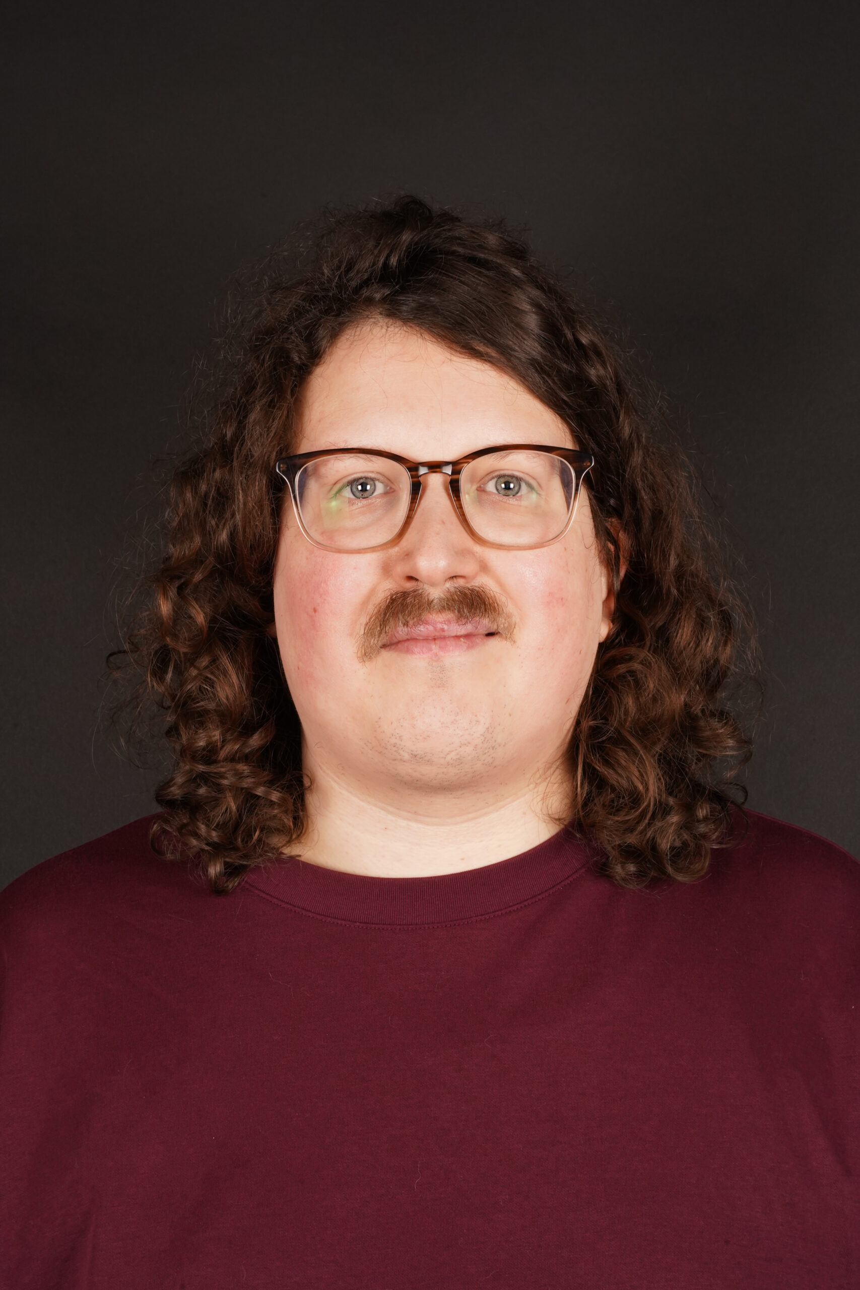 Portretfoto van Sem Bannenberg. Sem is een witte man van 35 met schouderlange, donkere krullen en een snor. Hij draagt een bril en een rood shirt.